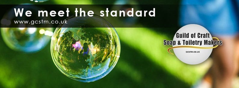 May include: A green grassy background with two soap bubbles. The larger bubble reflects a green tree and a blurred image of a person. The text "We meet the standard" and "www.gcstm.co.uk" is at the top of the image. A white circle with the text "Guild of Craft Soap & Toiletry Makers" and "gcstm.co.uk" is in the lower right corner.