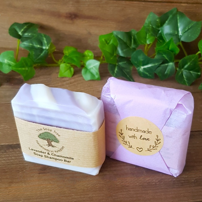 May include: A lavender and chamomile soap shampoo bar wrapped in brown paper with a label that reads "The Soap Tree Handmade in Yorkshire Lavender & Chamomile Soap Shampoo Bar". The bar is next to a purple gift bag with a brown circle sticker that reads "handmade with love".