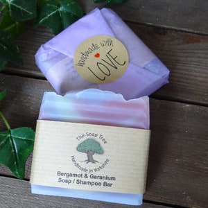 May include: A bar of Bergamot & Geranium soap/shampoo, wrapped in a paper sleeve and a lavender-colored paper. The soap has pink and white layers. A round sticker with "handmade with LOVE" is on top. The Soap Tree logo is visible.