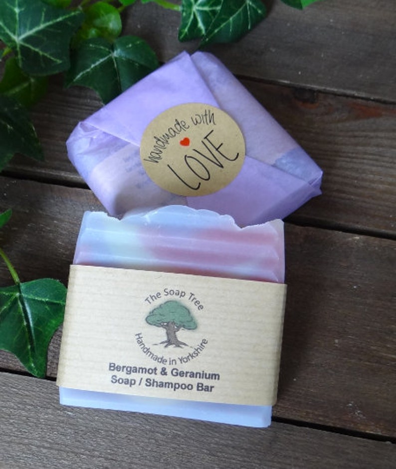 May include: A bar of Bergamot & Geranium soap/shampoo, wrapped in a paper sleeve and a lavender-colored paper. The soap has pink and white layers. A round sticker with "handmade with LOVE" is on top. The Soap Tree logo is visible.