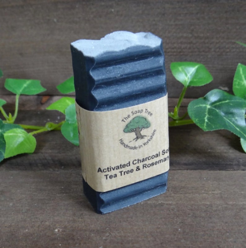 Puede incluir: Una pastilla de jab&oacute;n de carb&oacute;n negro rectangular con un borde superior texturizado. Una etiqueta de papel color canela envuelve el centro, con un logotipo de &aacute;rbol y el texto: The Soap Tree, Handmade in Yorkshire, Activated Charcoal Soap, Tea Tree & Rosemary.