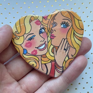 May include: Heart-shaped pin featuring a retro illustration of two women with blonde hair, blue eyes, and rosy cheeks. One woman is kissing the other's cheek. The pin has a vintage aesthetic with bold lines and bright colors.