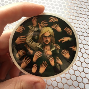 Op de afbeelding: Een sticker met een persoon met lang blond haar, omringd door veel handen die naar hen reiken. De persoon draagt ​​een donkergroen shirt en een witte kraag. De sticker heeft de tekst "Protect Your" onderaan.