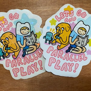 May include: Two cartoon characters, one with a yellow dog and the other with a blue shirt, are playing together. The text on the sticker says "Let's go and parallel play!"