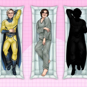 May include: Three rectangular body pillows with illustrations of figures. The first pillow features a figure in a yellow and blue superhero costume. The second pillow shows a figure in grey pyjamas. The third pillow displays a black silhouette of a figure in a superhero costume.