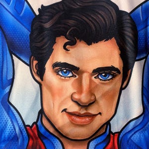 May include: Close-up of a portrait of a man with dark hair and bright blue eyes. He is wearing a blue and red costume. The man has a confident expression, with his arms behind his head.