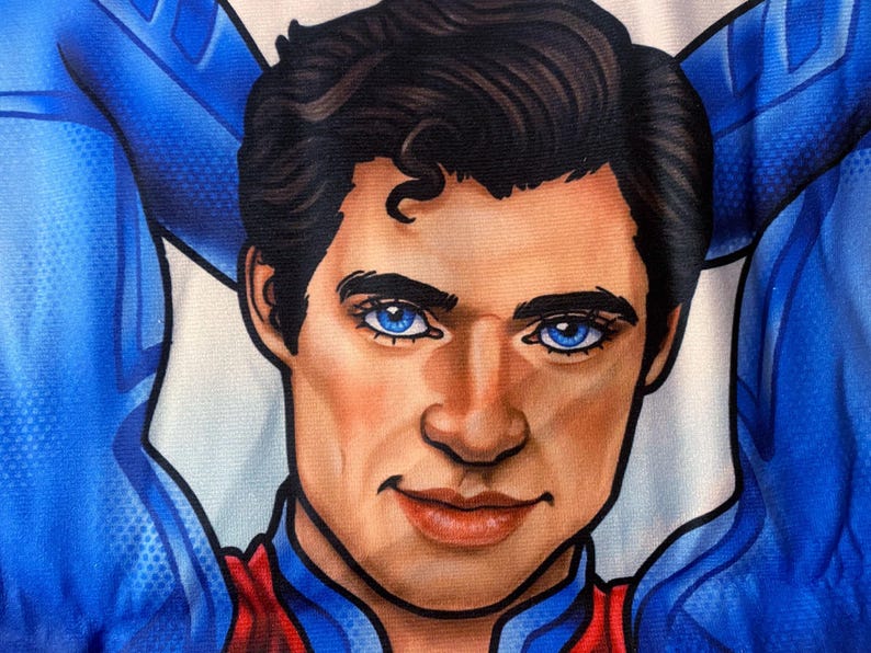 May include: Close-up of a portrait of a man with dark hair and bright blue eyes. He is wearing a blue and red costume. The man has a confident expression, with his arms behind his head.