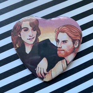 May include: Heart-shaped button featuring an illustration of two people with brown and red hair, set against a sunset-colored background. The button has a glossy finish and is placed on a black and white striped surface.