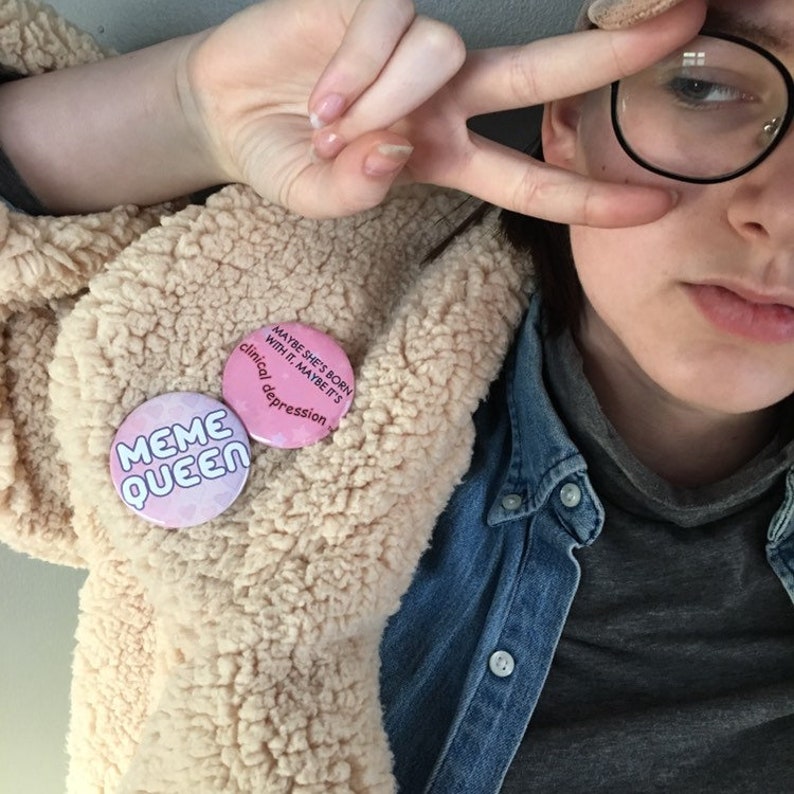 Puede incluir: Una persona que lleva una chaqueta de pelo beige, una camisa vaquera azul y un jersey de cuello alto gris. Lleva un bot&oacute;n rosa con el texto "Meme Queen" y otro bot&oacute;n rosa con el texto "Maybe she's born with it. Maybe it's clinical depression."