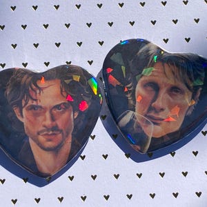 Może przedstawiać: Dwie ozdobne przypinki w kształcie serca z portretami dwóch mężczyzn. Przypinki mają holograficzny, błyszczący efekt. Tło jest białe z wzorem małych czarnych serc.