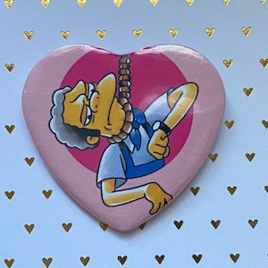 May include: A heart-shaped button featuring a cartoon character with gray hair and a blue shirt, appearing to hang from a noose. The button has a pink background with a pink circle in the center. The background is white with gold hearts.