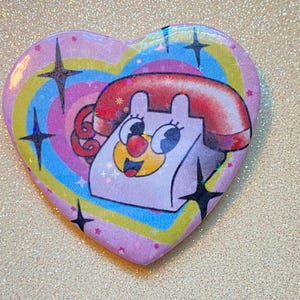 May include: Heart-shaped badge with a cartoon telephone design. The badge has a pink background with a rainbow swirl and black star accents. The telephone has a red receiver and a smiling face.