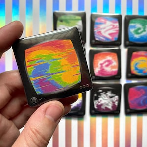 May include: A collection of square pins shaped like vintage television sets. Each pin features a black frame with a screen displaying a unique, colorful, abstract pattern with swirling or glitch-like designs in shades of pink, blue, yellow, and green.