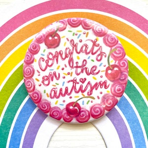 May include: A pink and white button with a cherry cake design and the text "Congrats on the autism" in pink lettering.