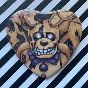 May include: Heart-shaped button featuring a detailed illustration of a character with a yellow-brown color scheme, a purple bow tie, and a sinister grin. The button is set against a black and white striped background.