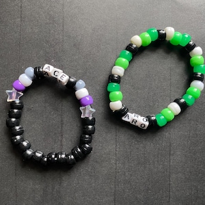 May include: Two beaded bracelets. One is black with white letter beads spelling "ACE", and purple and white beads with star accents. The other is green, white, and black beads with letter beads spelling "ARO".