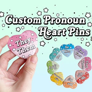 May include: A set of heart-shaped pins with different pronoun sets, including "They/Them", "Xe/Xim", "Fae/Faer", "It/Its", "She/Her", "He/Him", and "Any Pronouns". The pins are arranged in a circle, with the "They/Them" pin in the center.
