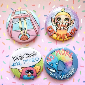 May include: Four round button pins with different designs. The first pin features a cartoon crab in a pink and white cage with the text "We the People are Pissed". The second pin features a cartoon character with a yellow face and red hair with the text "Eat the Rich". The third pin features a cartoon blue creature with the text "No More Billionaires". The fourth pin features a cartoon pink and blue creature with the text "We the People are Pissed".
