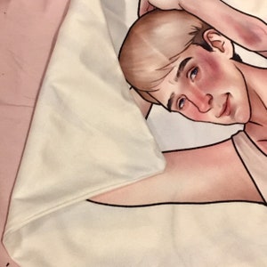 May include: A white fabric throw blanket featuring a printed illustration of a person with light brown hair, blue eyes, and flushed cheeks. The person is depicted in a reclining pose with one arm raised behind their head, rendered in a soft art style.