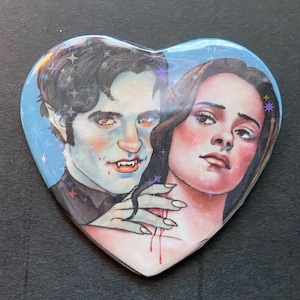 May include: Heart-shaped button with a vampire and a woman illustration. The vampire has pale skin, fangs, and dark hair. The woman has dark hair and a pale complexion. The button has a blue background with star accents and a glossy finish.