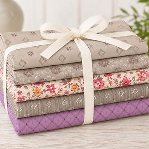 May include: A stack of five folded fabric pieces tied with a cream-colored ribbon. The fabrics feature various patterns including geometric diamonds, small floral sprigs, and larger pink and yellow floral prints in shades of beige, gray, and purple.