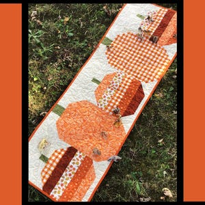 May include: A quilted table runner with orange pumpkin shapes, green stems, and a white background. The runner has a repeating pumpkin pattern and the text "Pumpkin Hollow". Designed by Tricia Lynn Maloney.