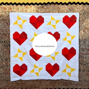 May include: White quilt featuring a repeating pattern of red hearts and yellow stars. The quilt is machine-stitched with a grid pattern. The text "VILLA ROSA DESIGNS" is in a white circle in the center of the quilt.