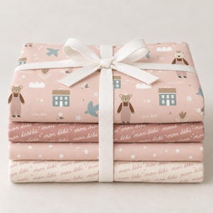 May include: A stack of four folded fabric pieces tied with a white ribbon. The top piece features a light pink background with illustrations of bears, houses, birds, and stars. The other three pieces feature a repeating script text reading mon bébé.