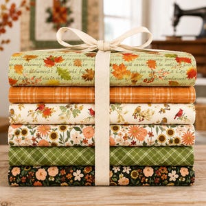 May include: A stack of six folded fabric pieces tied with a cream ribbon. The patterns include autumn leaves with cursive text, orange plaid, birds with foliage, orange and yellow flowers, green plaid, and a dark floral print with small flowers.