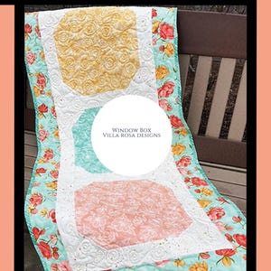May include: A quilted table runner with a floral pattern and three geometric shapes. The shapes are yellow, teal, and pink, with white floral detailing. The edges are a teal floral print. The text "WINDOW BOX VILLA ROSA DESIGNS" is in a white circle.