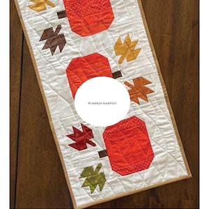 May include: A quilted table runner featuring a pumpkin and fall leaf design. The runner is primarily white with orange pumpkins and various colored leaves. The text "PUMPKIN HARVEST" is in the center. Finished size is 13" x 42".
