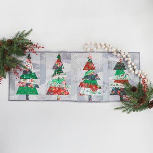 May include: A rectangular Christmas table runner featuring four stylized Christmas trees. The trees are made of fabric with red, green, and white floral patterns. The background is a patchwork of gray and white fabrics. Decorative greenery and a beaded garland frame the runner.