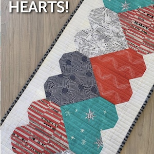 May include: A quilt table runner with a "Let's Play Hearts!" title. The runner is made of white fabric with a row of patchwork hearts in various patterns and colors, including red, blue, and teal.