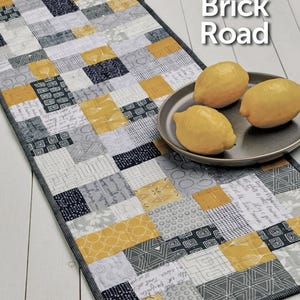 May include: A gray and yellow patchwork table runner with a black border. The runner is made of squares of different fabrics with various patterns. The runner is on a white wooden surface. The text "Mini Brick Road" and "ATK-205" are visible in the image.