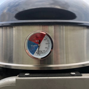 May include: Close-up of a black and silver barbecue grill with a temperature gauge. The gauge displays temperatures in Fahrenheit, with a blue section for smoking and a red section for grilling. The grill's lid is black and shiny.