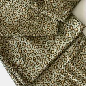 May include: A folded, decorative table runner with a leopard print pattern. The runner features a light green background with brown and tan spots. The edges are trimmed with a darker green border. The fabric appears to be a soft, silky material.