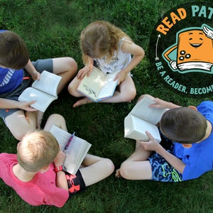 May include: Four children sit in a circle on green grass, each reading a book. A colorful logo in the background reads "Read Patrol: Be a reader. Become a leader."