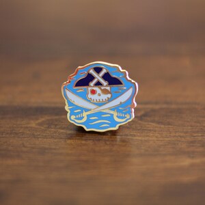 May include: Gold-colored enamel pin with a blue and white design. The design features a skull wearing a black and white pirate hat with crossed swords behind it. The skull has a red eye and white teeth.