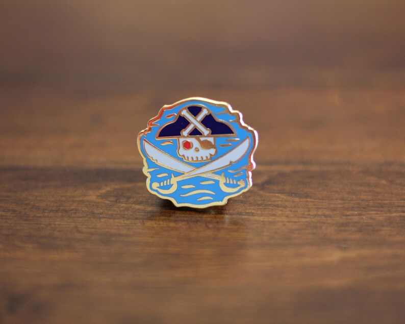 May include: Gold-colored enamel pin with a blue and white design. The design features a skull wearing a black and white pirate hat with crossed swords behind it. The skull has a red eye and white teeth.