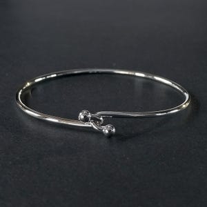 May include: A silver-colored bangle bracelet with a simple, elegant design. The bracelet features a slender, curved band that tapers to two small, round ball accents on one end. The other end has a hook closure. The jewelry is displayed against a dark background.