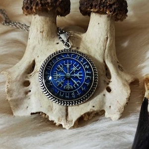 Könnte beinhalten: Eine silberne Halskette mit einem blauen Glasanhänger, der einen Kompass mit Runen am Rand zeigt. Der Anhänger ist von einem silbernen Rahmen mit einem Seilmuster umgeben.