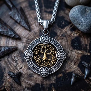Könnte beinhalten: Silberfarbene Halskette mit einem Anhänger, der ein goldfarbenes Lebensbaum-Design zeigt. Der runde Anhänger hat keltische Knotensymbole und Runen. Die Kette hängt an einer silbernen Kette. Der Hintergrund ist eine dunkle Holzoberfläche.