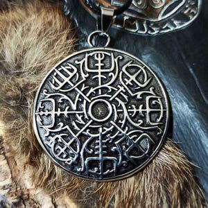 May include: A silver pendant with an elaborate circular design, displaying various symbols and patterns. It hangs from a black cord. The background features fur and a dark, textured surface, creating a rustic or historical aesthetic.