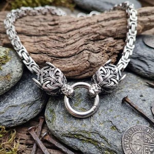 Könnte beinhalten: Eine silberfarbene Metallkettenhalskette mit einem dicken byzantinischen Gliederkettenmuster. Die Kette endet in zwei detaillierten, skulptierten Tierkopf-Endstücken, die einen kreisförmigen Ringverschluss in der Mitte des Schmuckstücks halten.