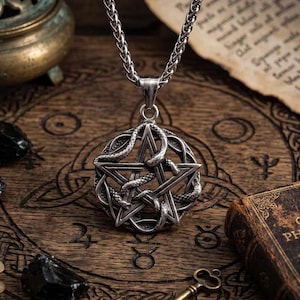 Könnte beinhalten: Eine silberfarbene Halskette mit einem Anhänger in Form eines Pentagramms, um das sich eine Schlange windet. Der Anhänger hängt an einer Kette. Die Kette liegt auf einer Holzoberfläche mit anderen okkulten Gegenständen.