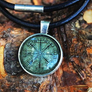 Könnte beinhalten: Eine silberne Halskette mit einem runden Anhänger, der einen grünen Kompass mit Runen zeigt. Der Anhänger ist an einem schwarzen Lederband.