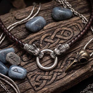 Könnte beinhalten: Eine braune Lederkette mit silbernen Drachenkopf-Akzenten und einem silbernen Verschluss. Die Halskette wird auf einer Holzoberfläche mit blauen Steinen und silbernen Kettendetails präsentiert. Ein Anhänger in Form eines bronzenen Vogelschädels ist ebenfalls vorhanden.