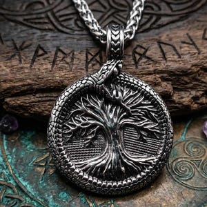 May include: A silver pendant necklace with a detailed tree design within a circular frame, encircled by a snake. The pendant hangs from a silver chain. The background includes wooden and metallic surfaces with engraved patterns and runes.