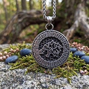 Könnte beinhalten: Halskette mit silberfarbenem Anhänger. Der runde Anhänger zeigt einen Wolf und Runen. Die Kette ist silberfarben. Die Kette liegt auf einem moosigen Felsen mit kleinen Steinen. Der Anhänger hat einen Durchmesser von ca. 3 cm.