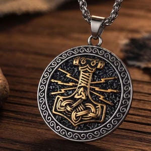 Könnte beinhalten: Ein kreisförmiger silberfarbener Anhänger mit einem goldfarbenen Mjolnir-Hammer-Design in der Mitte, umgeben von Blitzmotiven auf einem strukturierten schwarzen Hintergrund. Der Anhänger ist von einem silbernen Rand mit Spiralmuster umgeben.